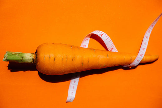 A fresh carrot wrapped in a measuring tape against a bright orange background