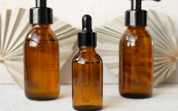 Three amber glass bottles with pumps and dropper set on a minimalist backdrop